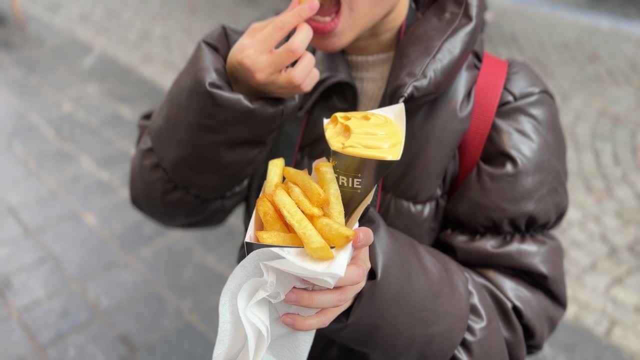 Girl eating Belgian fries with mayonnaise served in a paper cone on a Brussels street