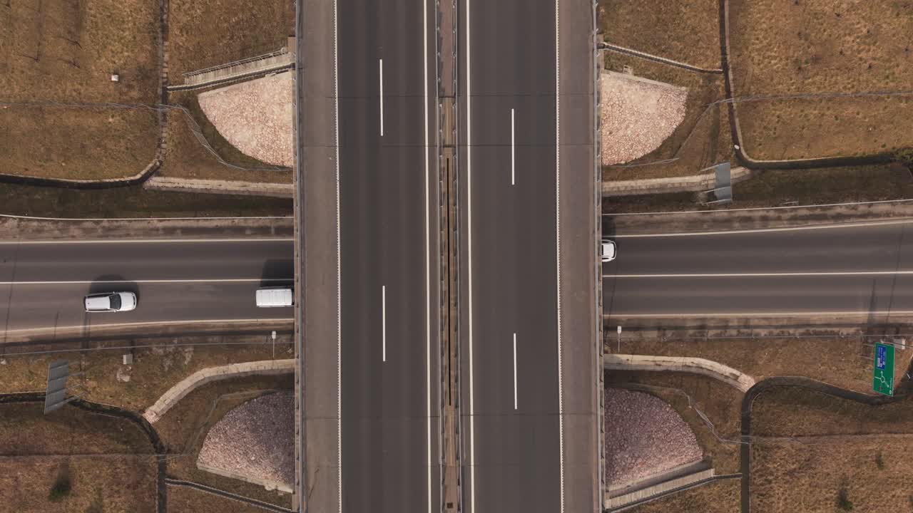 Top-down aerial drone view of a highway overpass where two roads intersect at different levels. Vehicles travel along the lanes while the surrounding rural landscape and agricultural fields frame the