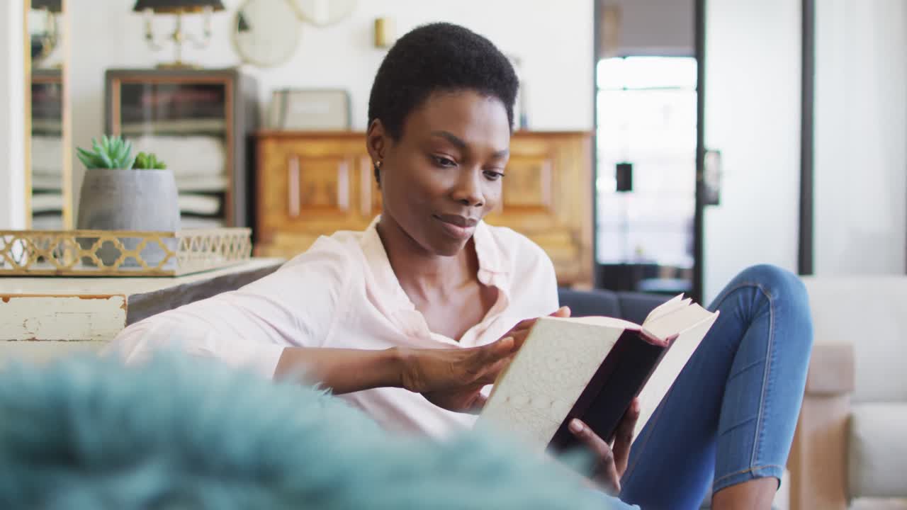 Glückliche afroamerikanische Frau sitzt auf dem Sofa im Wohnzimmer und liest ein Buch.