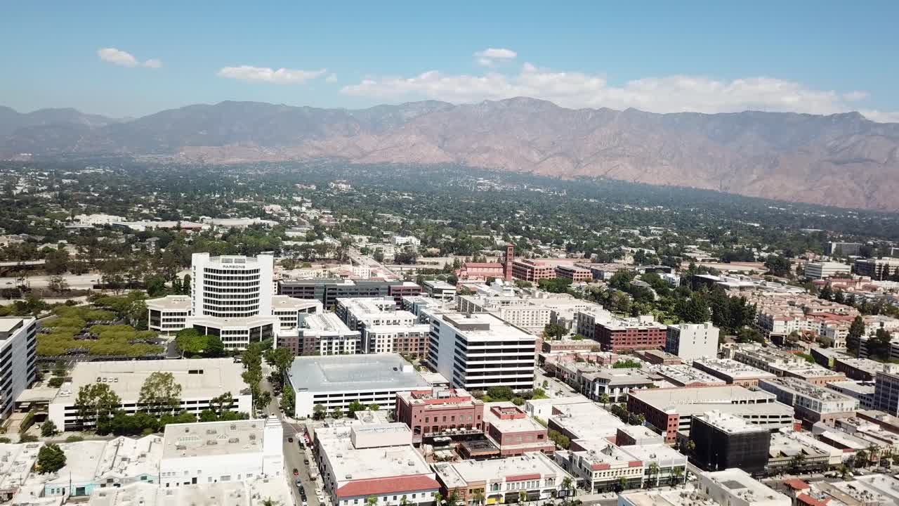 Vista aerea del centro di Pasadena, California, che mostra edifici cittadini, isolati residenziali, strade alberate e le montagne di San Gabriel sullo sfondo, in pieno giorno.