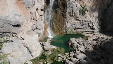 Still-steady-shot-slowly-zooming-out-as-freshwater-drops-from-a-cliff-at-a-bottom-of-a-waterfall,-forming-a-pond-by-carving-a-lake-in-the-rock,-an-exotic-green-pond