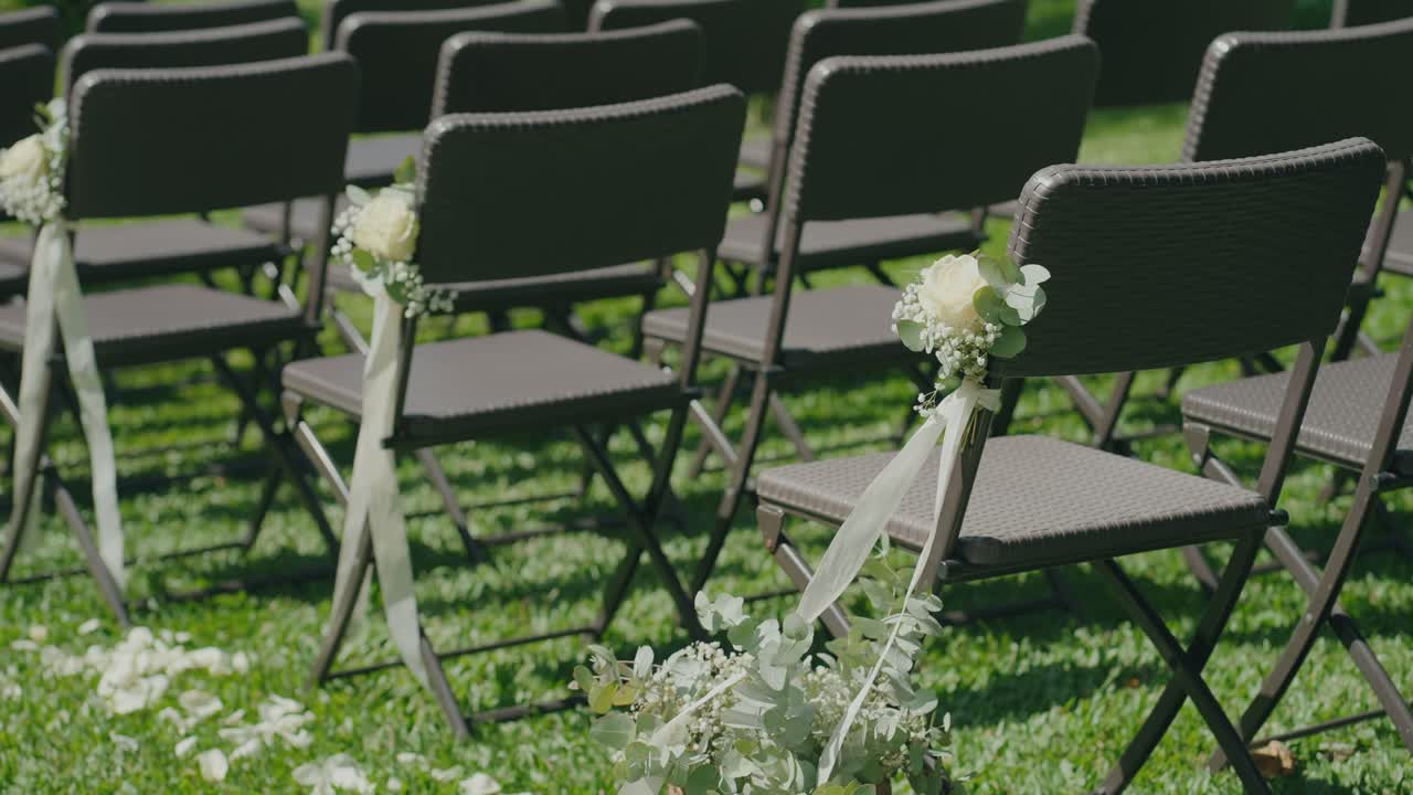 Rows of chairs decorated with white flowers and ribbons set up for an outdoor wedding ceremony on green grass