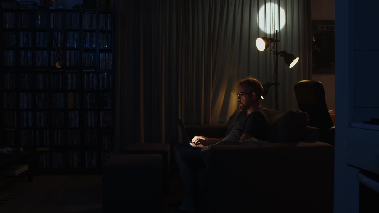 Zoom in to a man working on a laptop in his living room late at night