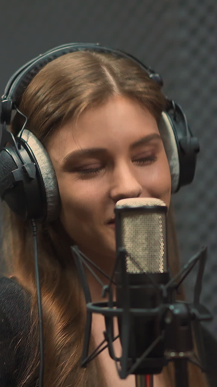 Attractive blonde long haired young woman with large headphones sings popular song with contemporary microphone in recording studio close view