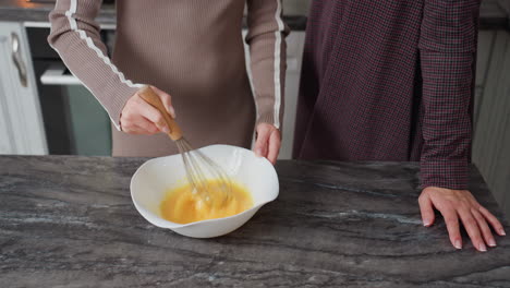 close-up of hands whisking eggs in white bowl on kitchen countertop, with observer nearby, kitchen scene captures food preparation, teamwork, and home cooking in warm domestic setting