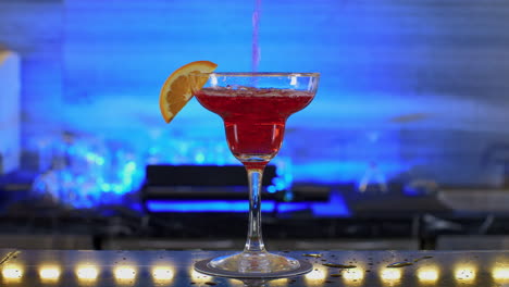 Rotes-Getränk-Füllt-Einen-Cocktailglasbecher-Mit-Orangenscheibe-In-Einem-Schicken-Nachtclub-Mit-Blauem-Licht-Im-Hintergrund