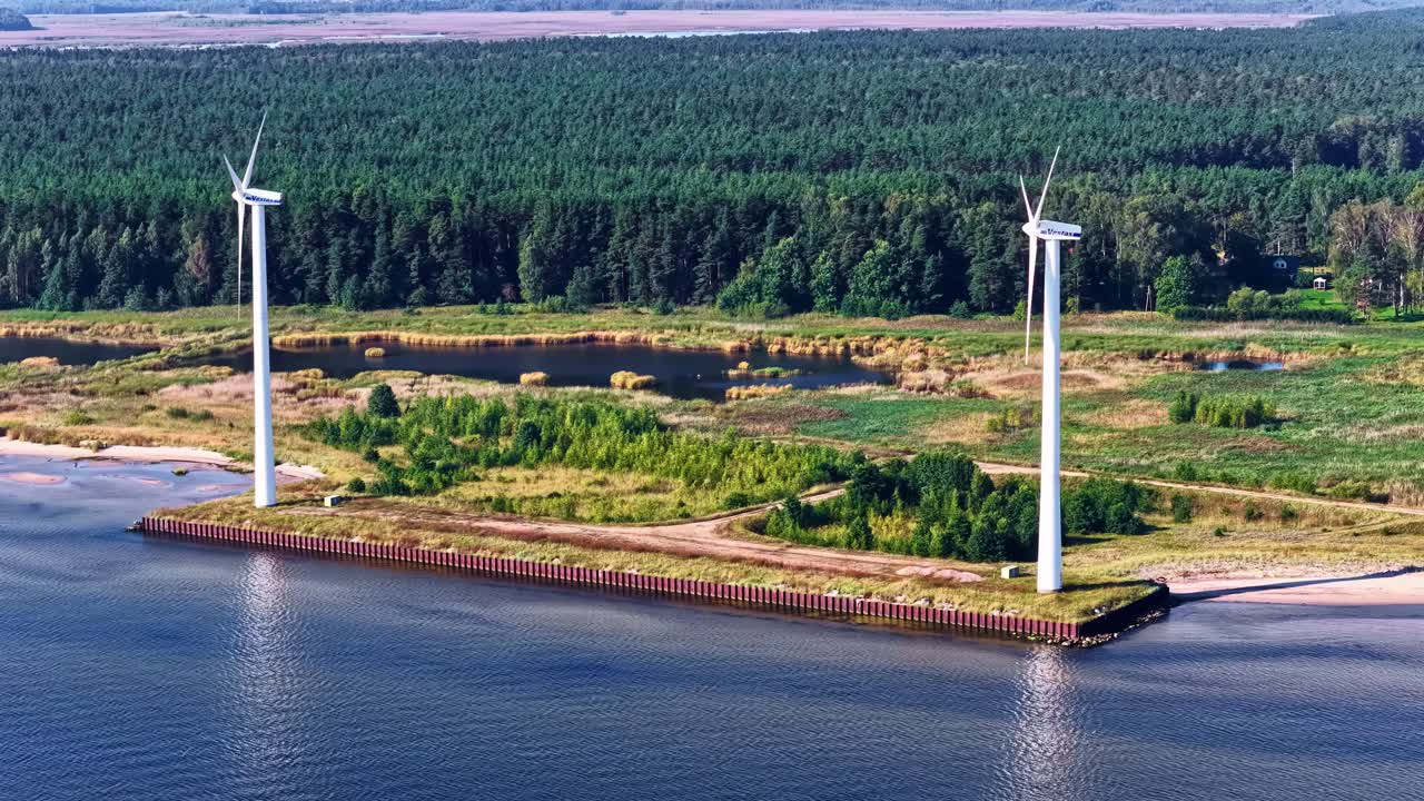 Two windmills on the bank of a body of water at the edge of a huge forest