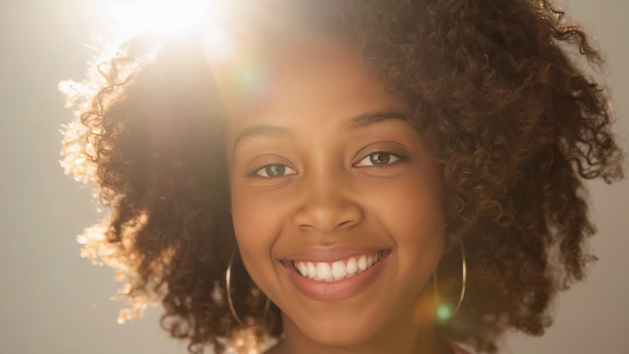 Modelo sorrindo e segurando o olhar enquanto a câmera faz zoom em um retrato de estúdio, com argolas, blusa rosa e luz de fundo