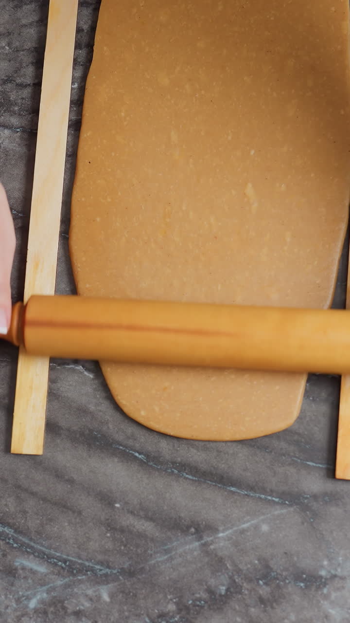 vista aérea de una mujer rodando masa con un rodillo de madera en la encimera, manos presionando firmemente, pulsera de cuentas rosas en la muñeca, rodeada de herramientas de horneado, cortadores de galletas, bandeja de madera y harina