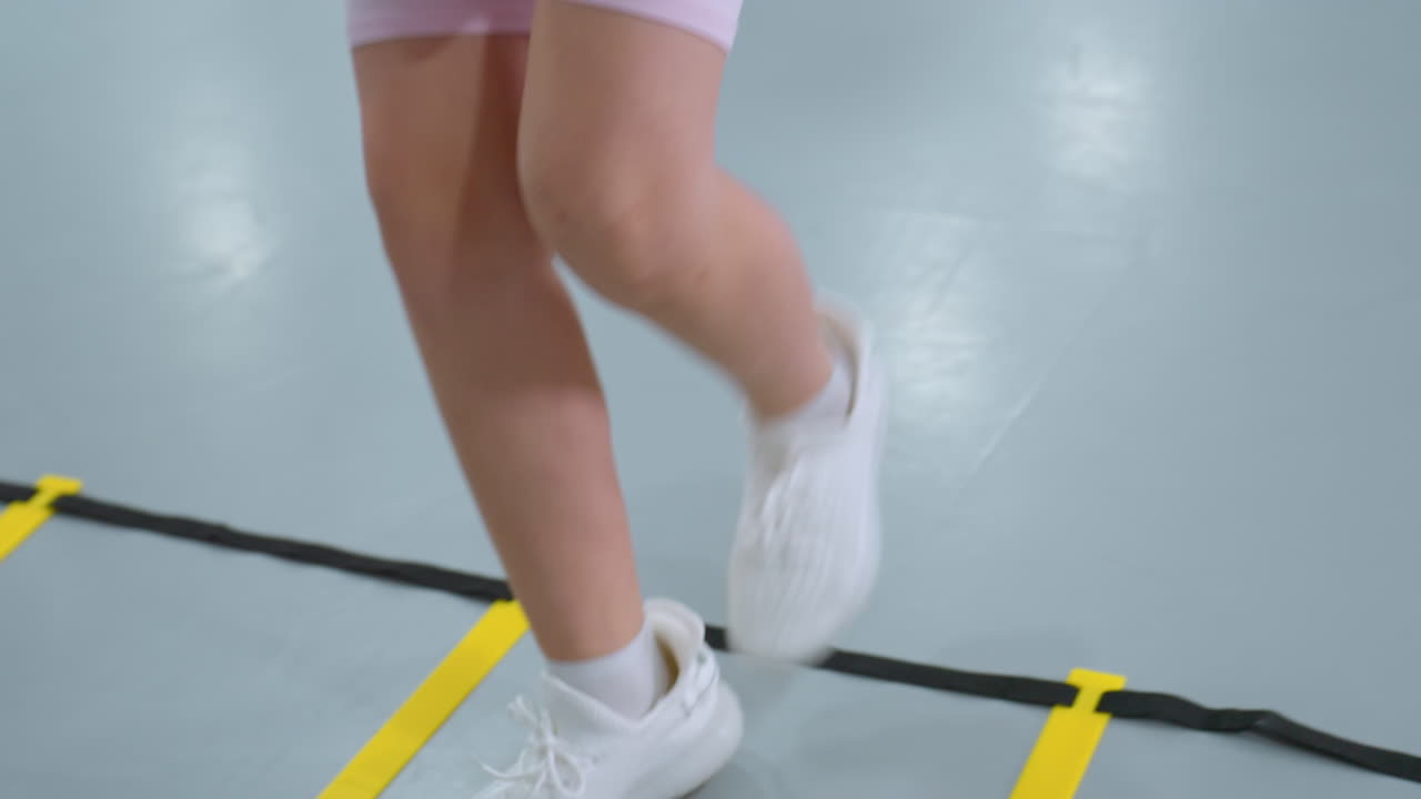 분홍색 셔츠를 입은 아이가 피트니스 운동을 하며 상자 안과 밖으로 발걸음을 옮기면서 다른 아이가 배경, 체육관 매트, 운동 액세서리 및 구조화된 피트니스 환경에서 훈련에 초점을 맞추고 있습니다.