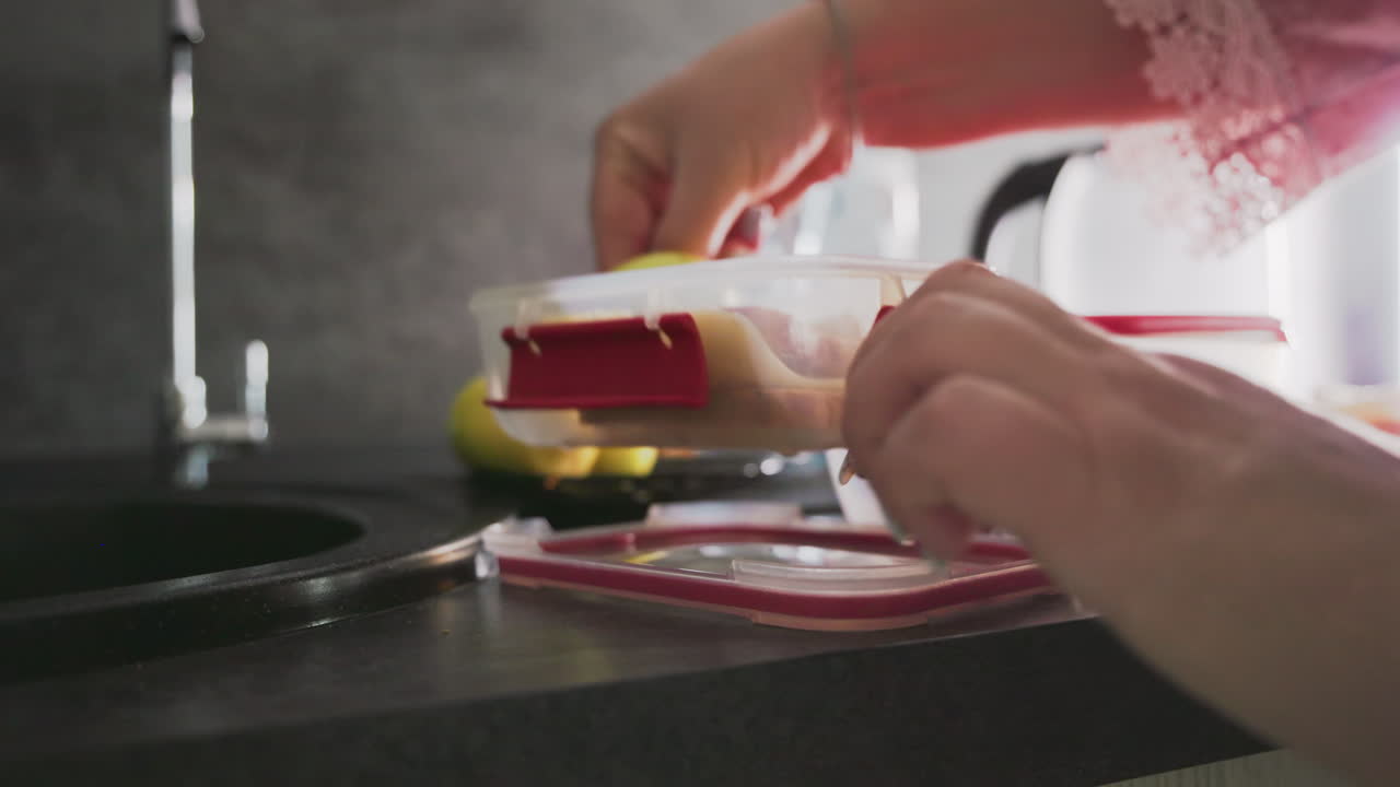 closeup of hand placing sandwich slices into plastic container with red lid on kitchen counter next to sliced bread, green apples, and faucet in bright light preparing healthy school lunch