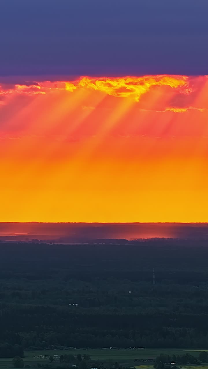 Intense orange and red sunset band light illuminates rays glowing above dark forest horizon, vertical timelapse