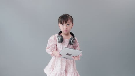 헤드폰을 끼고 있는 작은 아시아 소녀는 태블릿에서 온라인으로 비디오를 보고 행복하게 춤을 춥니다.