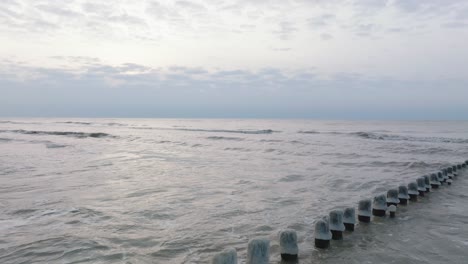 Luftaufnahme-Eines-Alten-Hölzernen-Piers-An-Der-Ostseeküste,-Bewölkter-Wintertag,-Weißer-Sandstrand-Mit-Schnee-Bedeckt,-Eis-Auf-Holzpfählen,-Ruhige-Küste,-Breiter-Drohnenschuss,-Der-Sich-Tief-Zurückbewegt