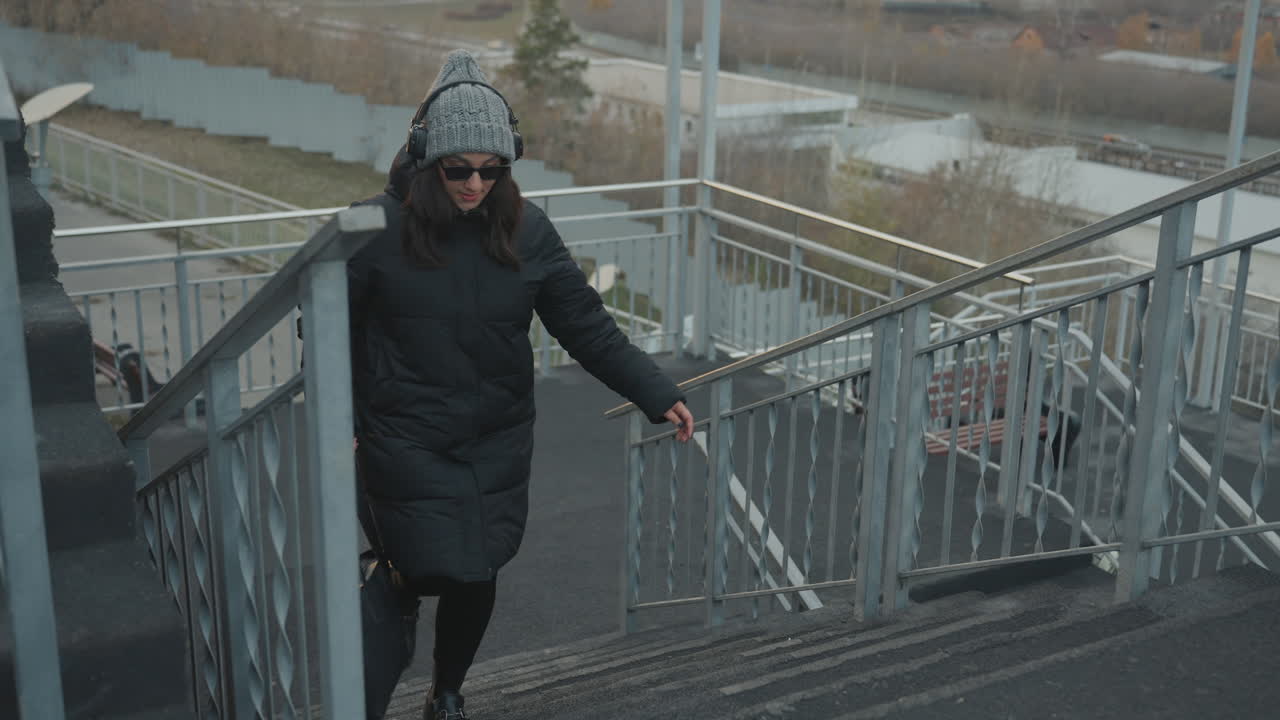 Woman walking up outdoor stairs wearing winter coat and beanie, carrying bag, listening to music and moving with subtle dance steps, metal railing and city skyline in soft dusk light, reflective