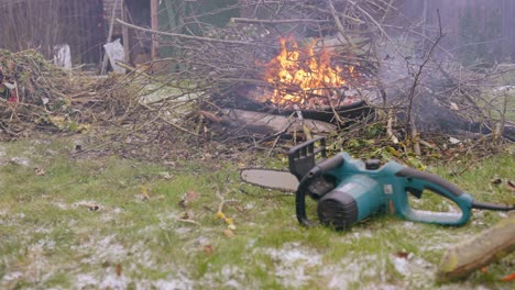 Person,-Die-Getrocknete-Zweige-In-Der-Natur-Am-Lagerfeuer-Platziert,-Kettensäge-In-Der-Nähe-Des-Feuers