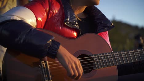 young female with shiny jacket playing guitar at sunset outdoors in mountains slow motion 60fps