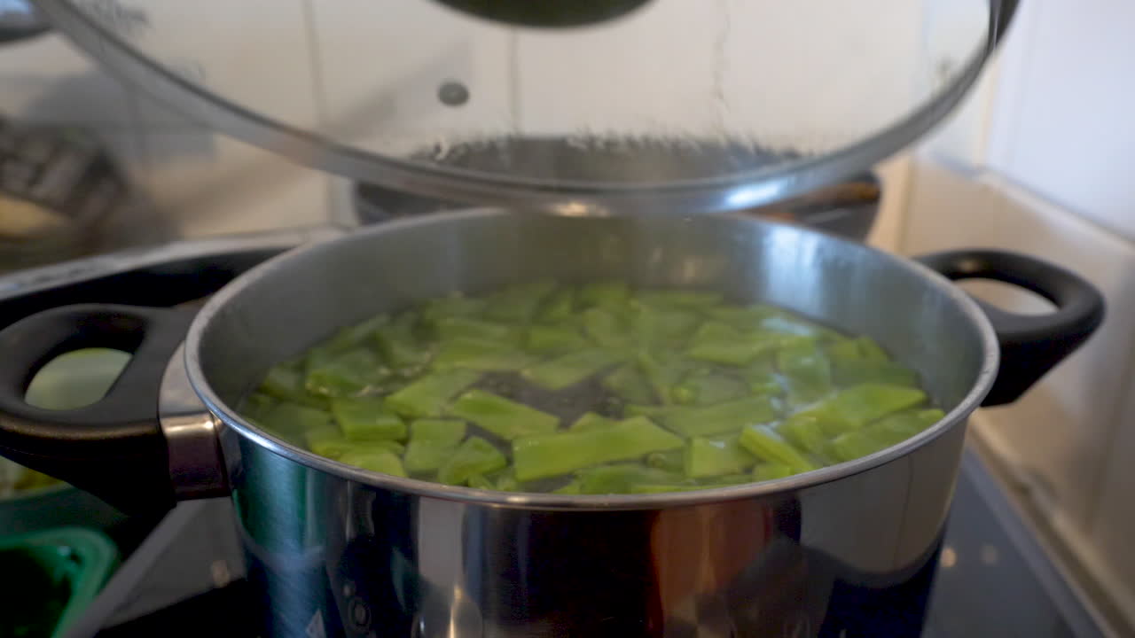 opening a lid of a bowl with cooking string beans in boiling water on top of a stove in a kitchen.