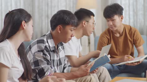 close up of asian teen group studying at home. a boy in plaid shirt with a book teaching a girl and frends, a boy holding a laptop discussing with his friends, writing into notebook.