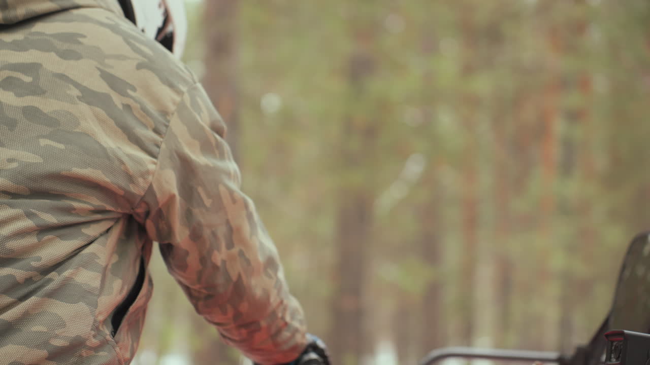 Un pilota caucasico afferra i comandi di un quad rosso con la mano guantata, primo piano del cruscotto e dell'acceleratore, giacca mimetica e casco visibili, sequenza di salita e accensione del motore in una pineta innevata, fuoristrada dinamico.