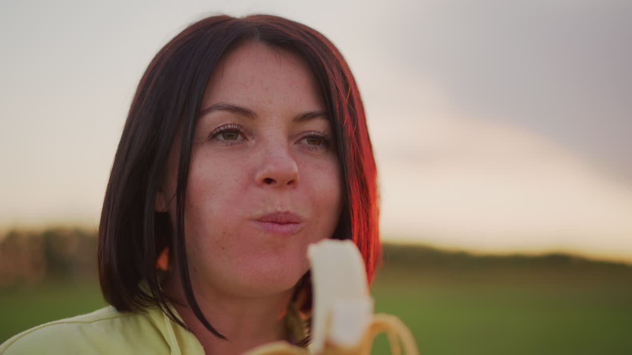 Caucasian woman eating banana during sunset postrun refresh in open field, neon jacket, playful smile while peeling banana, closeup portrait, warm rim light, relaxed breathing, healthy snack vibe