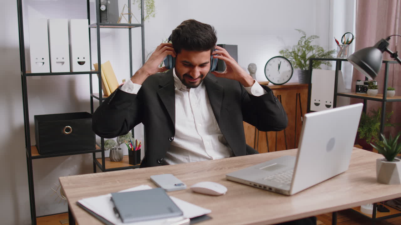 Indian businessman working on laptop at home office wearing headphones listening favorite music