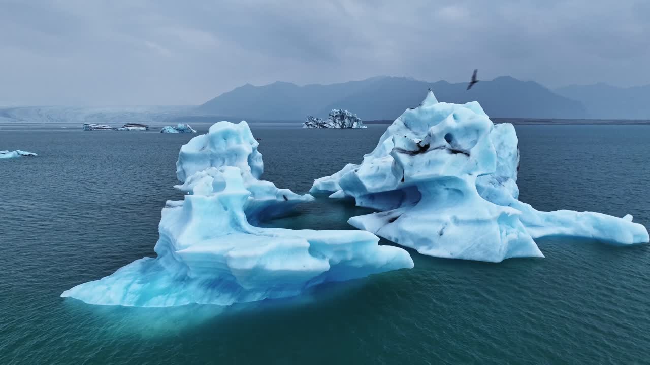 A smooth fly-through between drifting Vatnajokull icebergs reveals deep blue textures, cold Arctic water, and distant mountains under a muted Icelandic sky