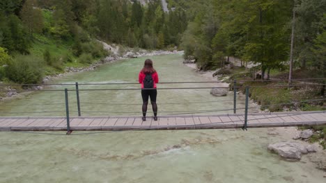 Young-woman-standing-on-suspension-bridge-above-a-calm-river-at-Soca-valley,-aerial