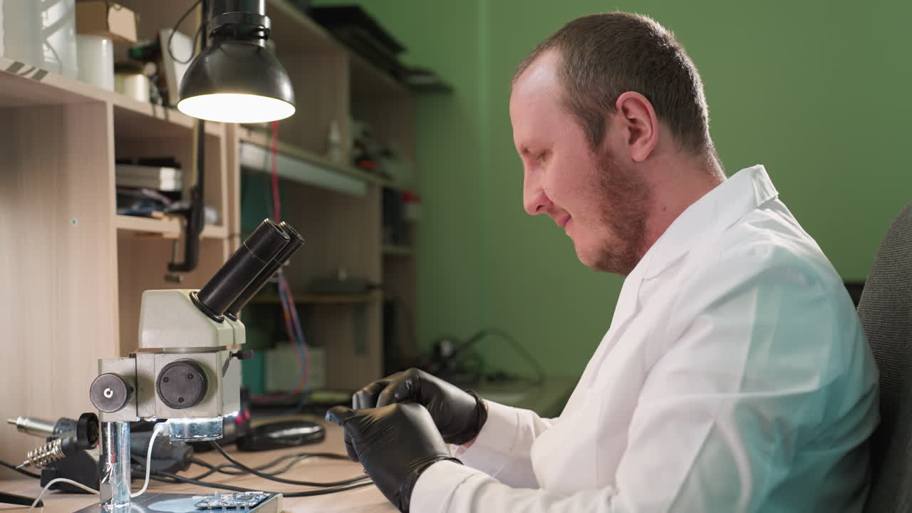 A middle view of a man wearing a white lab coat inside his lab removed his glasses and dropped the circuit under a microscope the laboratory setting includes a microscope and various tools