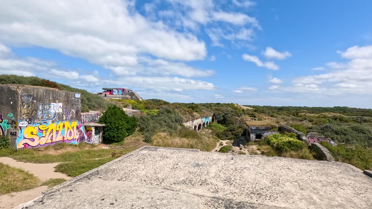 Camera pans across historic graffiti-covered concrete bunker amid greenery under bright, partly cloudy skies