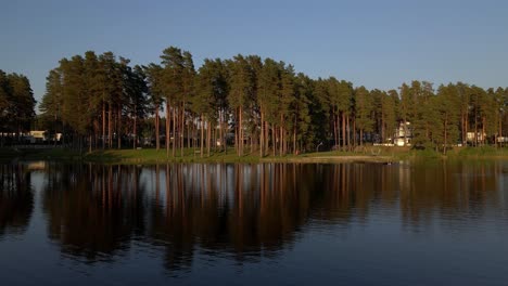 drone glides to the right over calm lake water near shore. trees line the lake with people swimming in the evening light.