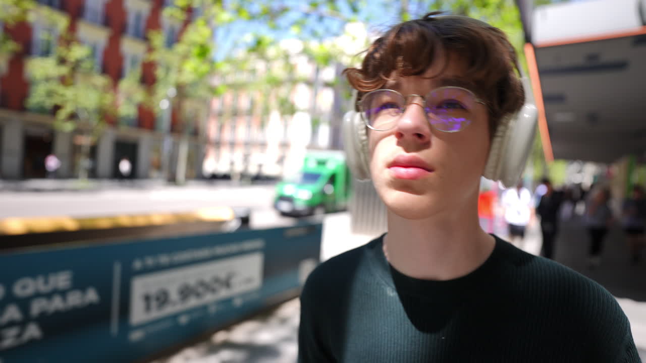 Teenager wearing headphones walking along sunny Madrid street, glancing around with genuine curiosity while experiencing urban lifestyle and enjoying musical moment