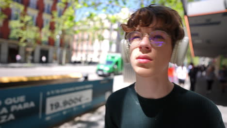 teenager wearing headphones walking along sunny madrid street, glancing around with genuine curiosity while experiencing urban lifestyle and enjoying musical moment