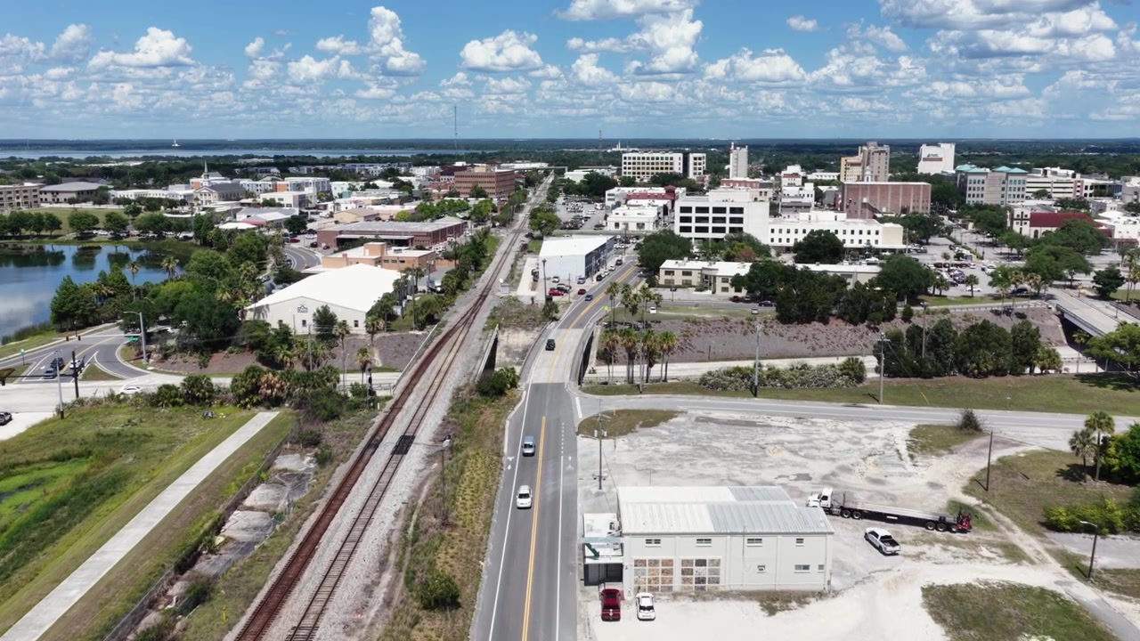 Biler kjører på delt motorvei ved siden av jernbanespor som fører mot Lakelands høyhus i sentrum, rolige innsjøvann glitrer under en delvis skyet Florida-himmel. Droneoverflyvning.