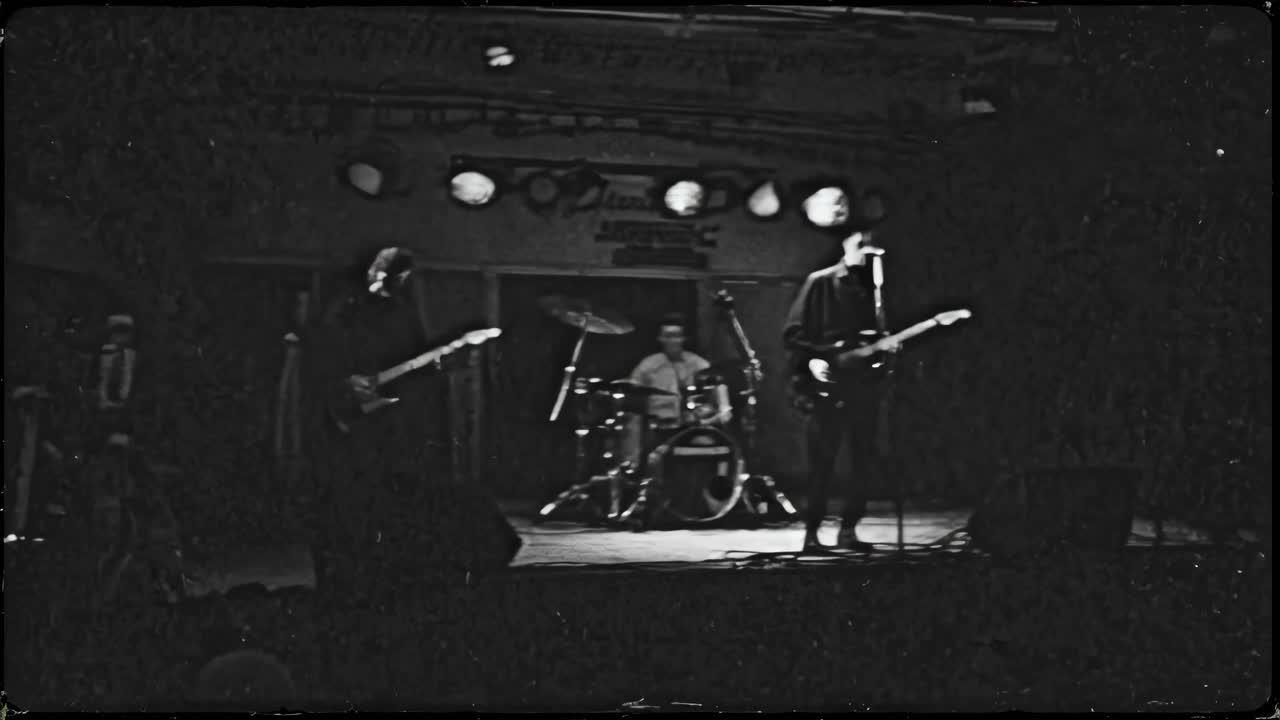 Grainy black-and-white video still of a band performing on stage, captured from a low angle