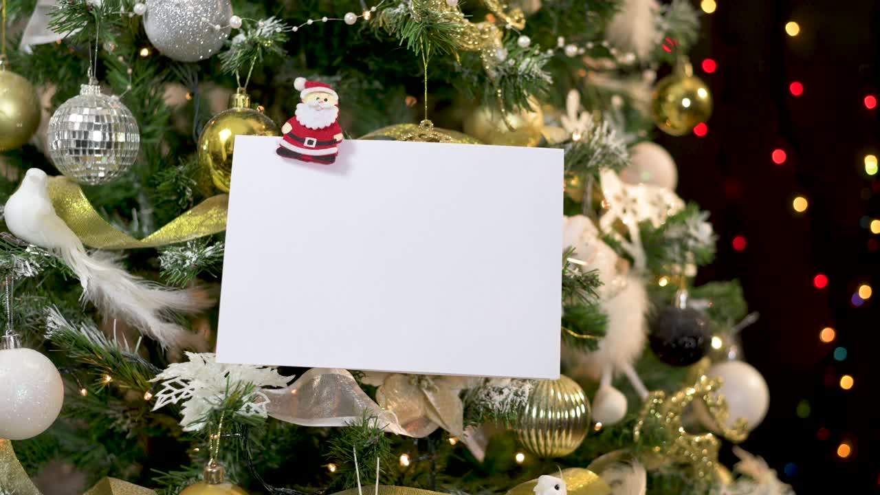 tarjeta de felicitación enganchada con ornamento de santa en oro árbol de navidad decorado en blanco con luces parpadeantes