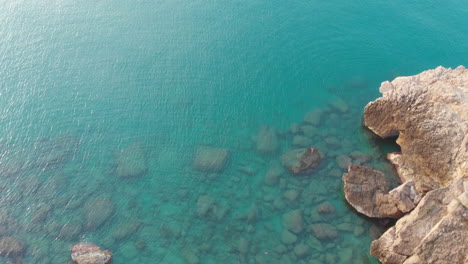 Vista-De-Pájaro-Capturada-Por-Un-Dron-Del-Borde-Del-Acantilado,-Mientras-La-Cámara-Se-Eleva-Sobre-Un-Paisaje-Escarpado-De-Agua-Azul-Y-Rocas-De-Arena-Blanca