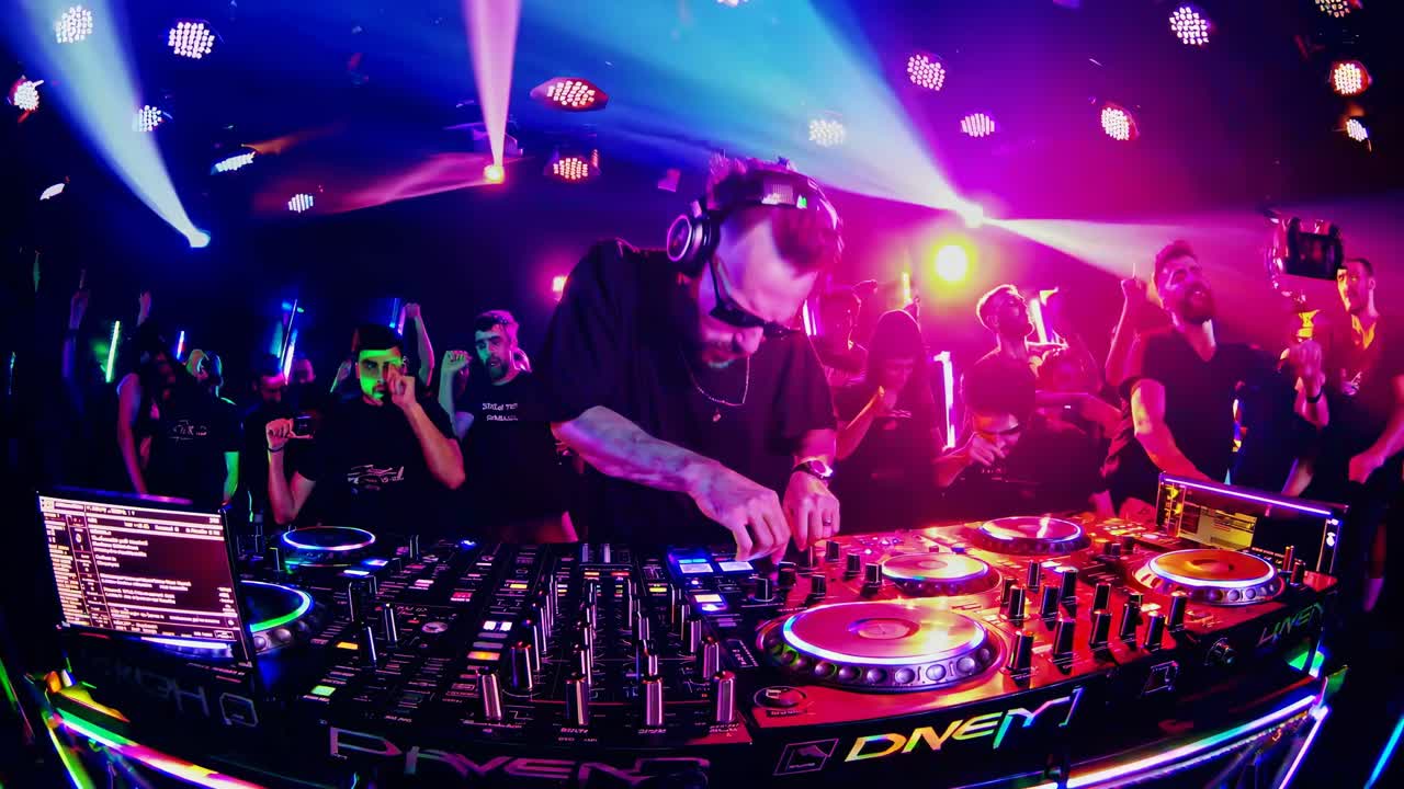 Plano de ângulo alto de um DJ em uma cena de boate vibrante com luzes coloridas e uma multidão animada.