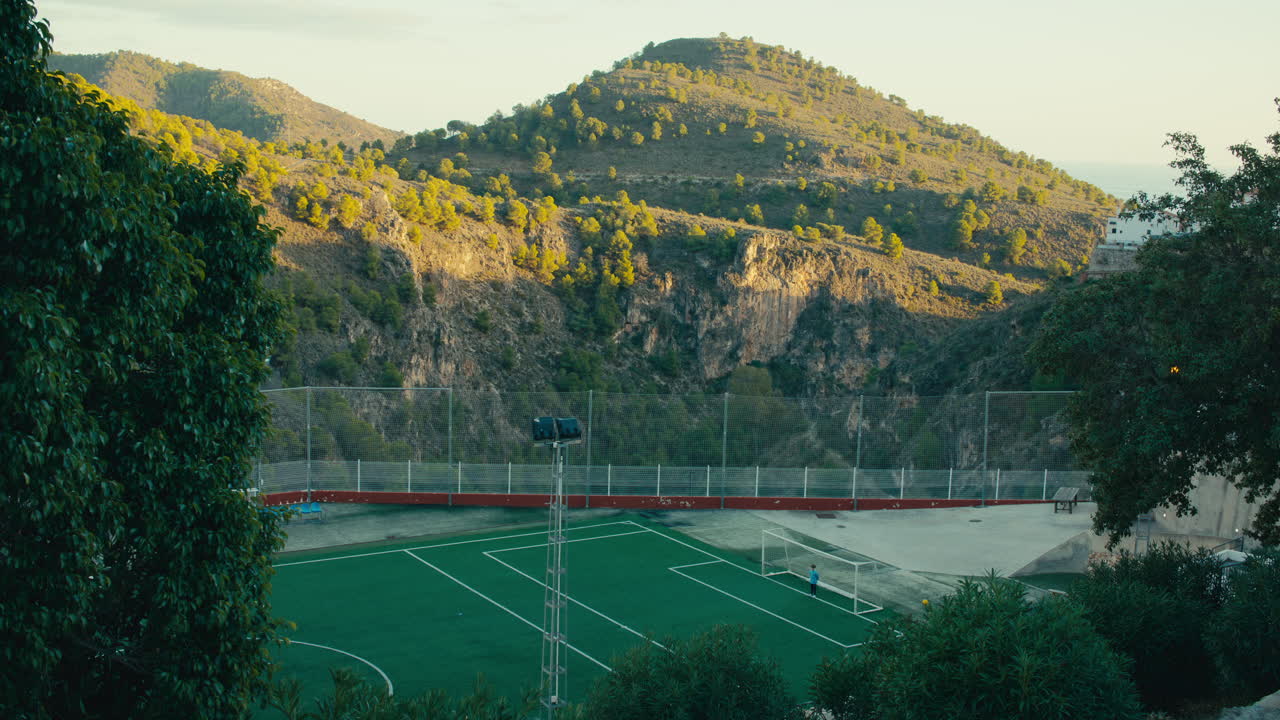 Kids practice football on a dramatic green field surrounded by cliffs and pine-covered hills in the Andalusian mountains