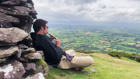 a young man playing a tune in a flute leaning on the rocks of an old castle in conor pass in ireland ovelooking green fields.