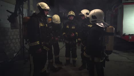 group of five firefighters wearing full equipment in smokey area. relaxed chatting after work well done. fire trucks in the background. name tag translation is fireman and model's last name.