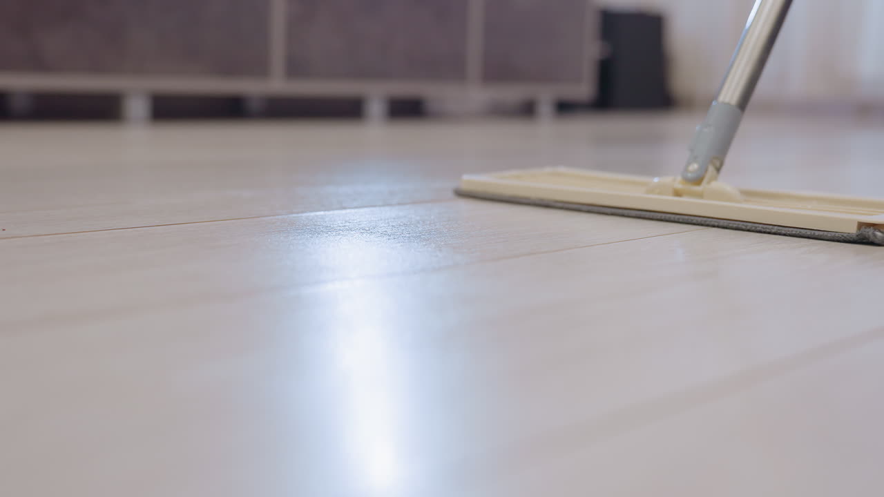 Close up of mop head being pushed across shiny wooden floor indoors showing reflection and smooth texture during household cleaning routine highlighting cleanliness