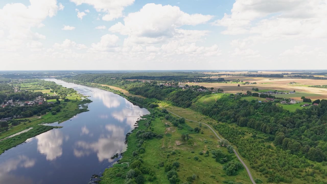 Splendide riprese aeree mostrano un fiume tranquillo che serpeggia attraverso una rigogliosa campagna verde, delimitato da campi e foreste. La luce solare intensa, il cielo blu e le nuvole soffici aggiungono un'atmosfera pacifica e naturale.