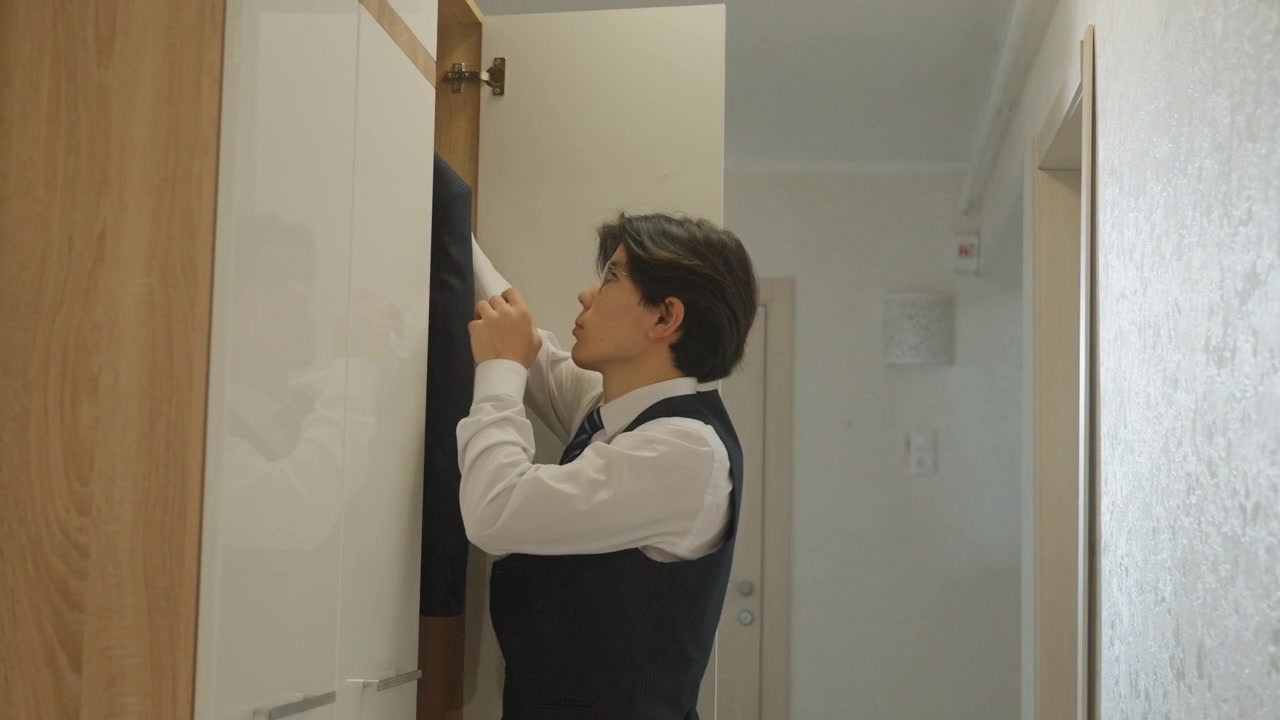 Giovane uomo asiatico che apre l'armadio, selezionando una giacca e un gilet blu navy, aggiustando polsini e bottoni, controllando la silhouette nello specchio, camminando nel corridoio di un appartamento moderno, atmosfera mattutina calma con luce solare calda.