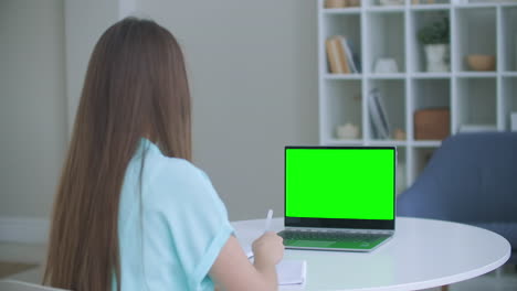 girl taking notes in notepad from laptop computer green mockup screen. girl learning online on computer