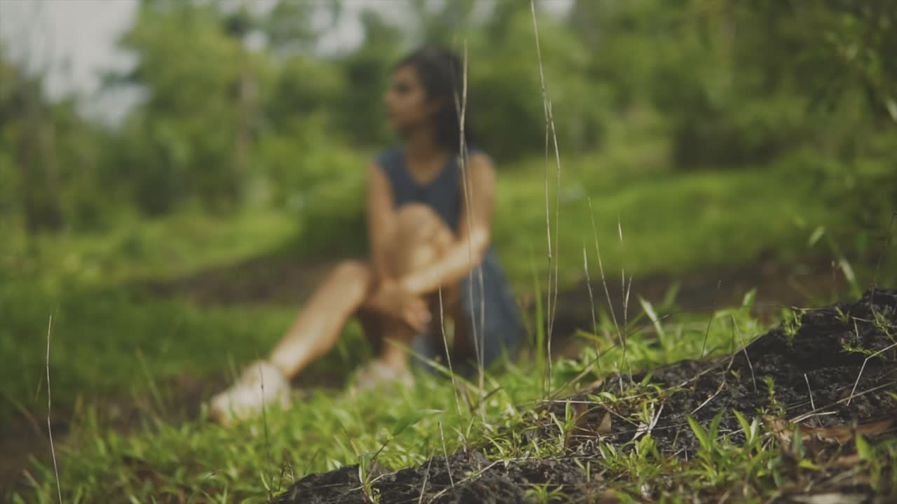 Tan girl sits on grassy hillside on sunny day pondering, thinking, focus on grass in front