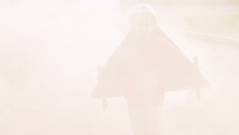 공원에서 달리고 조종사로 노는 뒤에 인공 연기가 있는 마분지 비행기 날개를 달고 있는 행복한 어린 소년
