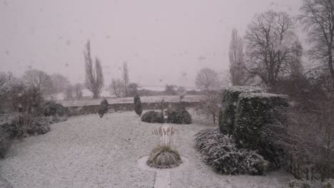 A serene snowy village garden in Northamptonshire, viewed through a window.