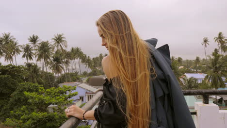 female model posing from behind walking along balcony with tropical background and throws clothing over camera lens