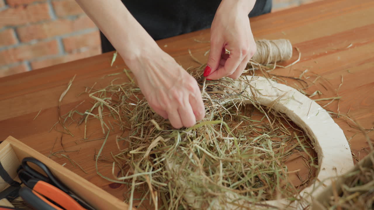 Decorator crafting rustic wreath with dried hay on circular base, using hands with red nails to carefully arrange material, with twine spool and tools nearby on wooden table in creative workspace